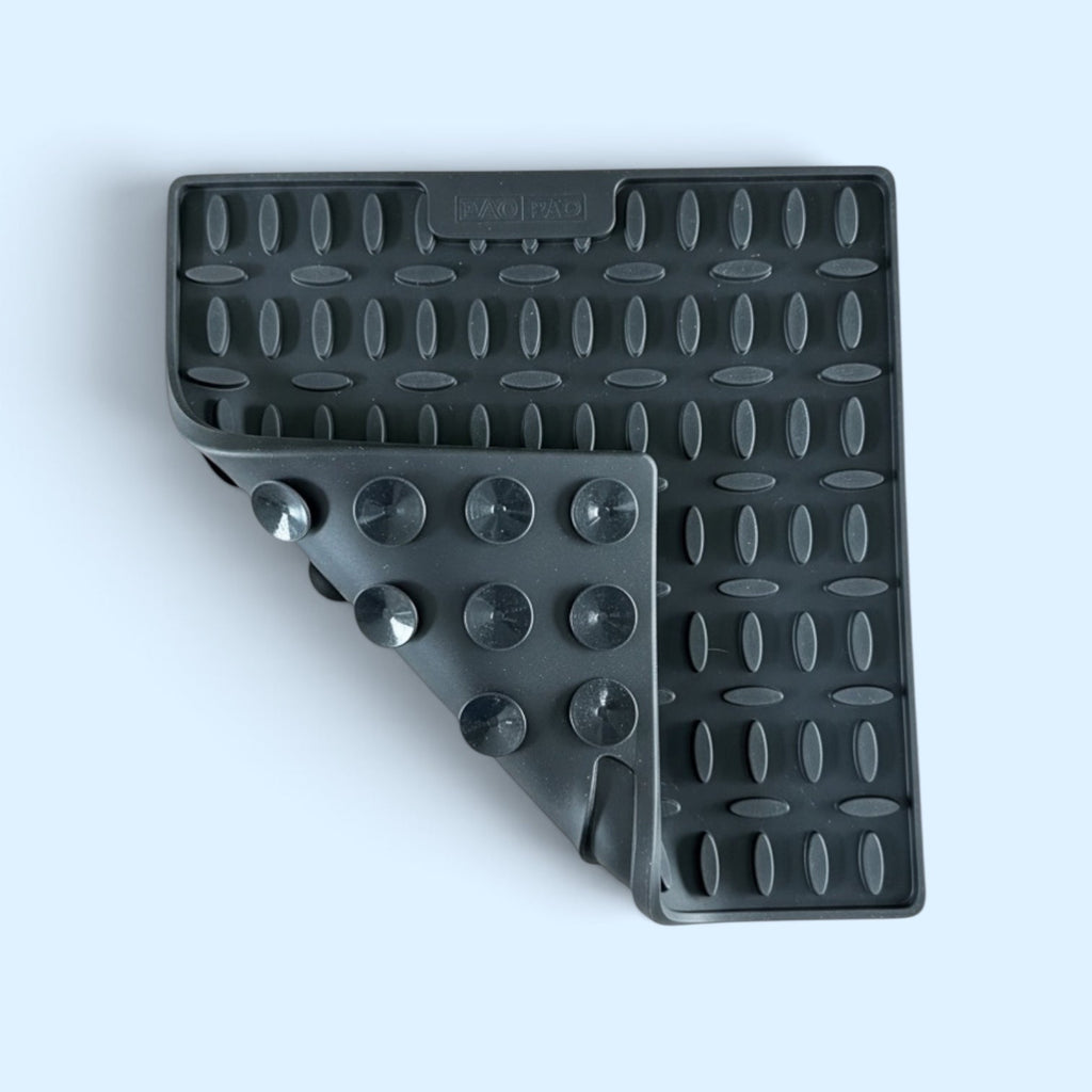 A black silicone square trivet with textured grip patterns, shown on a light blue background.