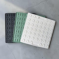Three square textured silicone trivets in black, mint green, and white, arranged diagonally on a gray concrete floor.