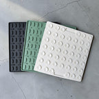 Three square textured silicone trivets in black, mint green, and white, arranged diagonally on a gray concrete floor.