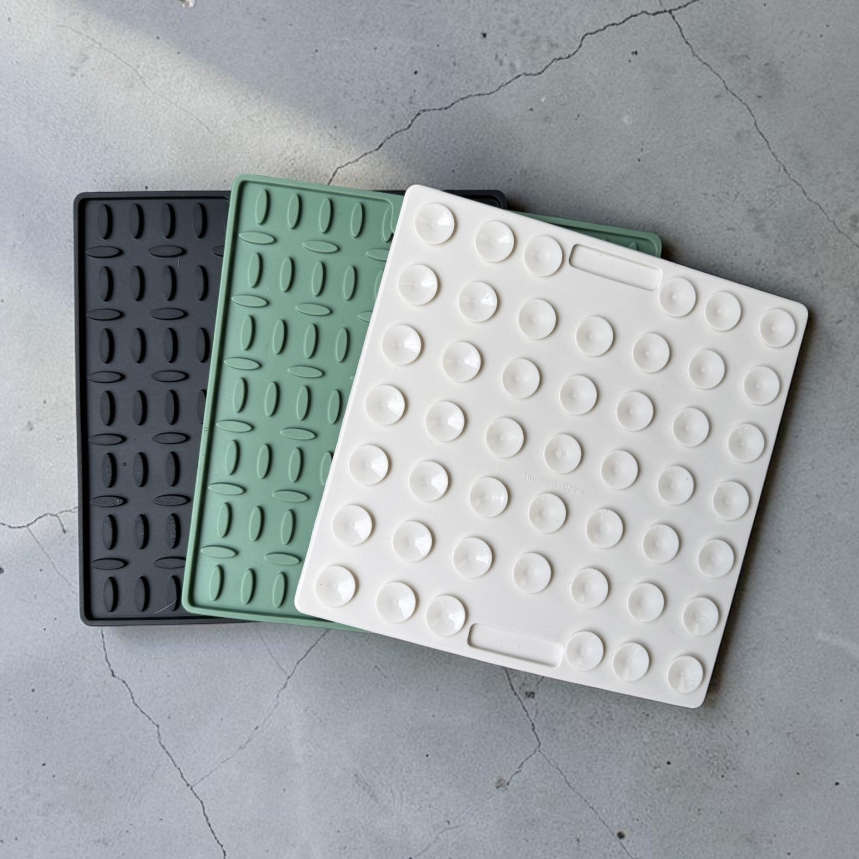 Three square textured silicone trivets in black, mint green, and white, arranged diagonally on a gray concrete floor.