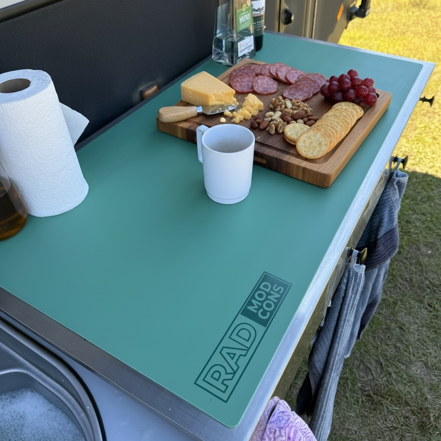 A eucalyptus coloured RAD MAT on the side of a camper van, styled with a charcuterie board, cheese, crackers, grapes, a mug, and a paper towel roll in an outdoor camping setting.