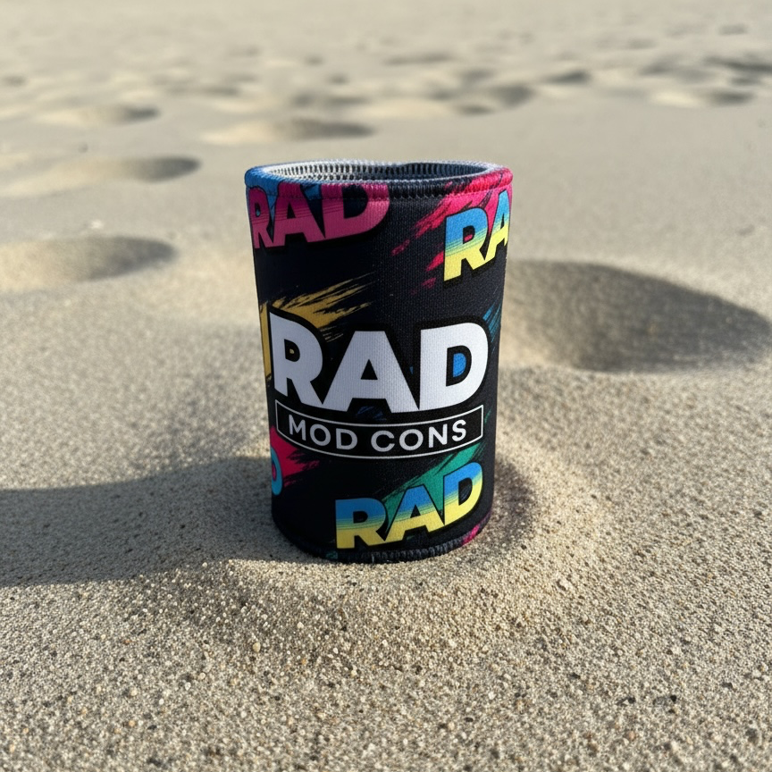 A black neoprene can cooler with a colorful graphic pattern and the text 'RAD MOD CONS' stands upright in a sandy beach setting.