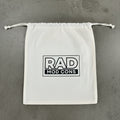 A white drawstring dust bag with a black 'RAD MOD CONS' logo printed on the front, laid flat on a gray concrete surface.