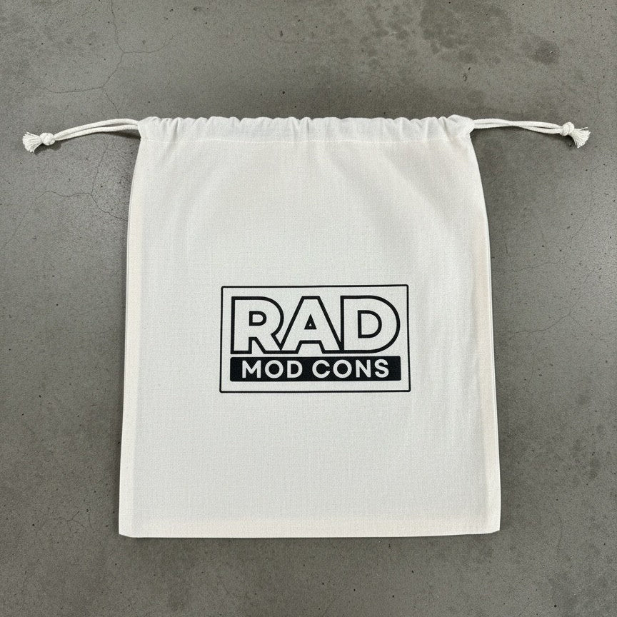 A white drawstring dust bag with a black 'RAD MOD CONS' logo printed on the front, laid flat on a gray concrete surface.