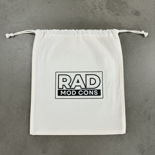 A white drawstring dust bag with a black 'RAD MOD CONS' logo printed on the front, laid flat on a gray concrete surface.